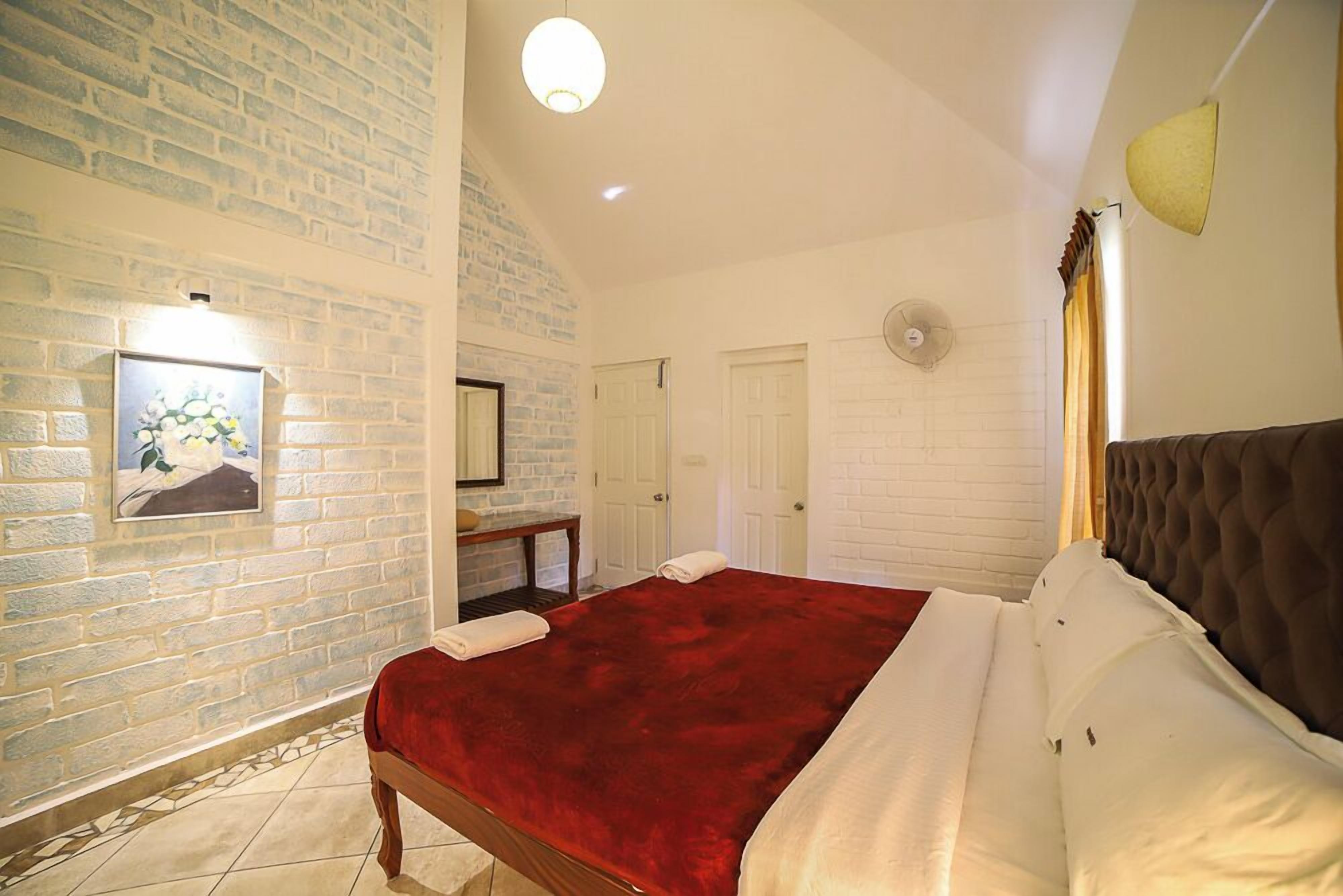 A bedroom with a brick wall, a bed with a red blanket, a wooden bench, and a picture on the wall.