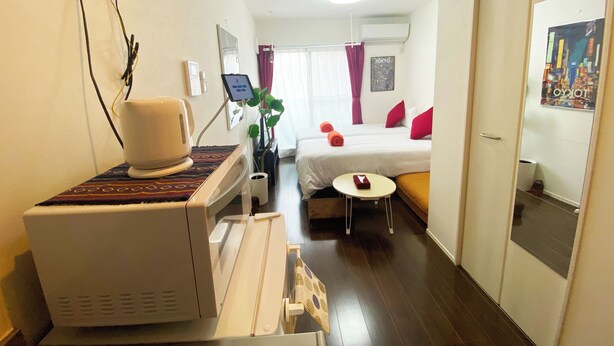A hotel room with a bed, bedside table, and a small seating area. There is a wall-mounted heater and a plant in the corner. - Skytree & Asakusa in front! Newly built clean apt! (墨田区)