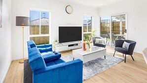 Una sala de estar moderna con un televisor de pantalla plana grande, un sofá azul, una mesa de centro con objetos decorativos y un comedor con mesa y sillas.