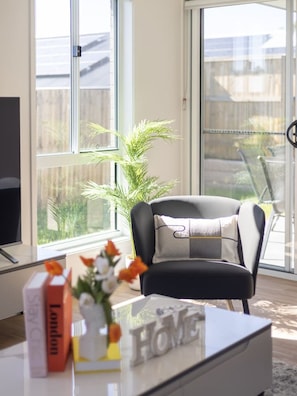 A modern living room with a black armchair, a glass coffee table, and a view of a backyard through large windows. - Brand New Comfy Home, 4Bedroom, designer furniture (Greenbank)