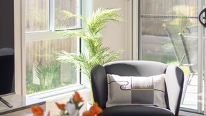 Una sala de estar moderna con un ventanal grande, un sillón negro con cojín, una mesa de centro de vidrio y una estantería con libros y un jarrón decorativo.