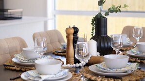 Mesa de comedor servida con platos, cuencos y copas de vino blancos, rodeada de sillas color beige.