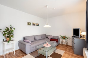 Un salon moderne avec un canapé gris, une table basse en verre, un téléviseur posé sur un meuble et un luminaire suspendu.