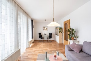 A modern living room with a sofa, a dining table with chairs, a hanging light fixture, and a potted plant.