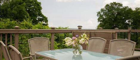 A glass-top table with wicker chairs on a balcony, surrounded by greenery and a clear sky.