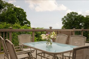 A glass-top table with wicker chairs on a balcony, surrounded by greenery and a clear sky.
