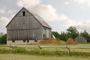 A large wooden barn with a stone foundation, hay bales, and a smaller adjacent barn in a rural setting.