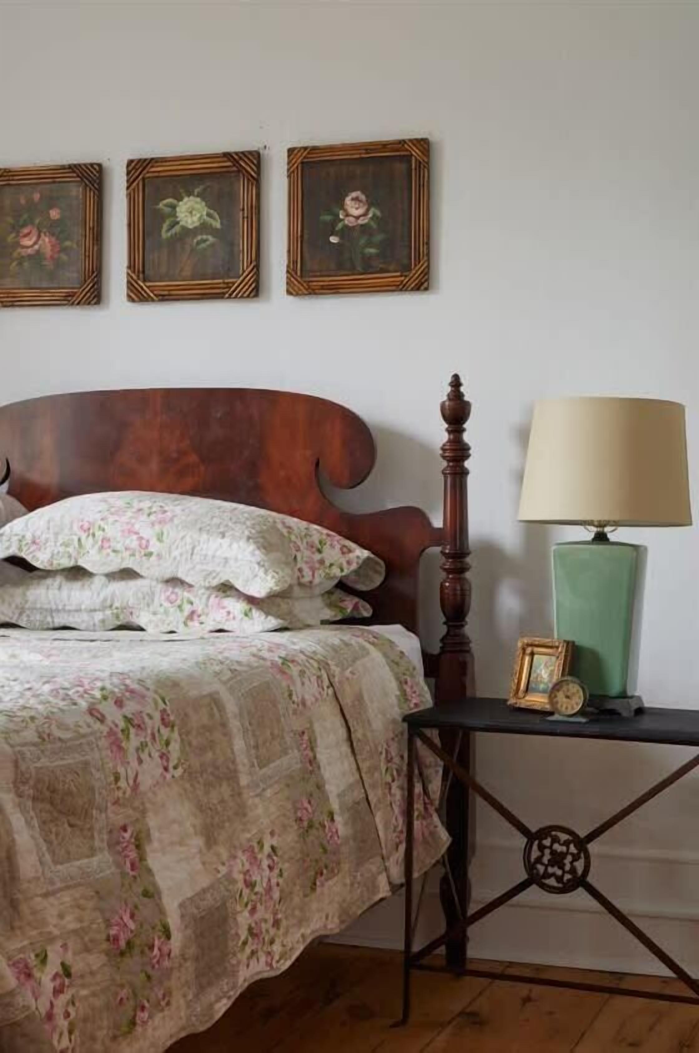 A bedroom with a wooden bed, floral bedding, a bedside table with a lamp, and three framed floral paintings on the wall.