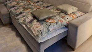 A bedroom with a sofa bed, a dining table with chairs, a floral-patterned bedspread, a grey sofa, a hanging light fixture, and a wall-mounted picture of a lynx. - Apartment Eschenweg incl. balcony with garden view, bed linen and towels (Bad Harzburg)