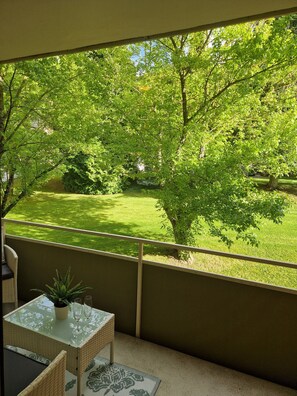 A balcony with a view of a green lawn and trees, a glass table with a potted plant, and wicker furniture.