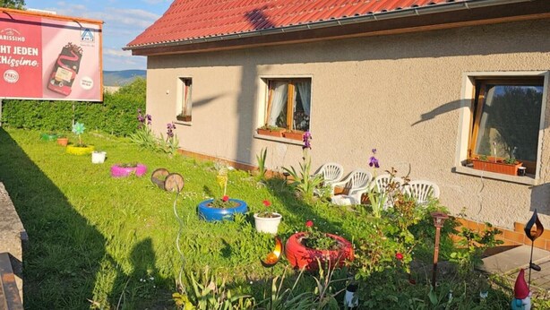 A house with a red-tiled roof, a garden with various plants and outdoor furniture, and a billboard in the background. - Apartment 'Ferienwohnung Peter' (Rudolstadt)