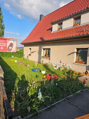 A house with a red-tiled roof, a garden with various plants and outdoor furniture, and a billboard in the background.