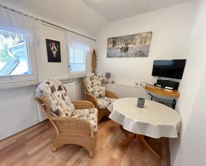 Una habitación acogedora con dos sillones de mimbre, una mesa redonda con mantel blanco, televisión y obras de arte en las paredes.