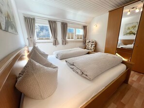 Un dormitorio con una cama, dos almohadas, una silla, una ventana con cortinas, un radiador y un espejo.