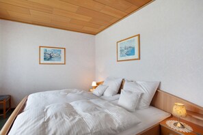 A neatly made bed with white linens, a wooden headboard, a bedside table with a lamp, and two framed pictures on the wall.