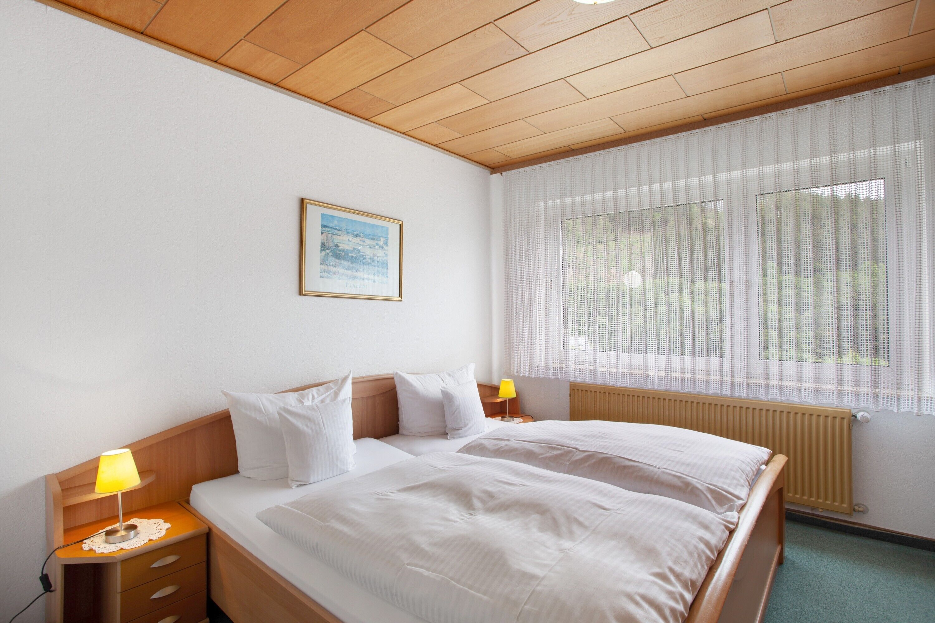 A hotel room with a wooden ceiling, a large bed with white bedding, a bedside table with a lamp, and a window with sheer curtains.