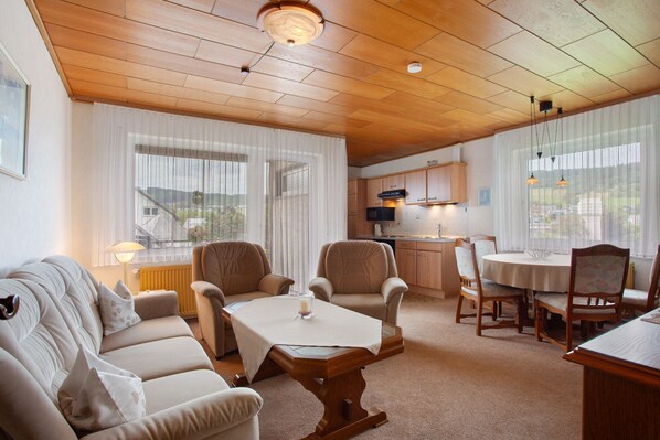 A cozy living room with a sofa, chairs, a dining table, and a kitchen in the background.