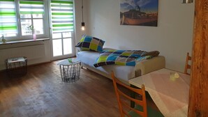 Ein gemütliches Zimmer mit einem Bett, einem Tisch und einem Stuhl. Ein Fenster mit Jalousien, eine Heizung und eine hängende Glühbirne sind ebenfalls vorhanden.