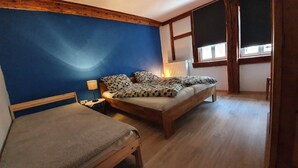 A bedroom with a bed, a smaller bed, a nightstand, a lamp, and a window with blinds.