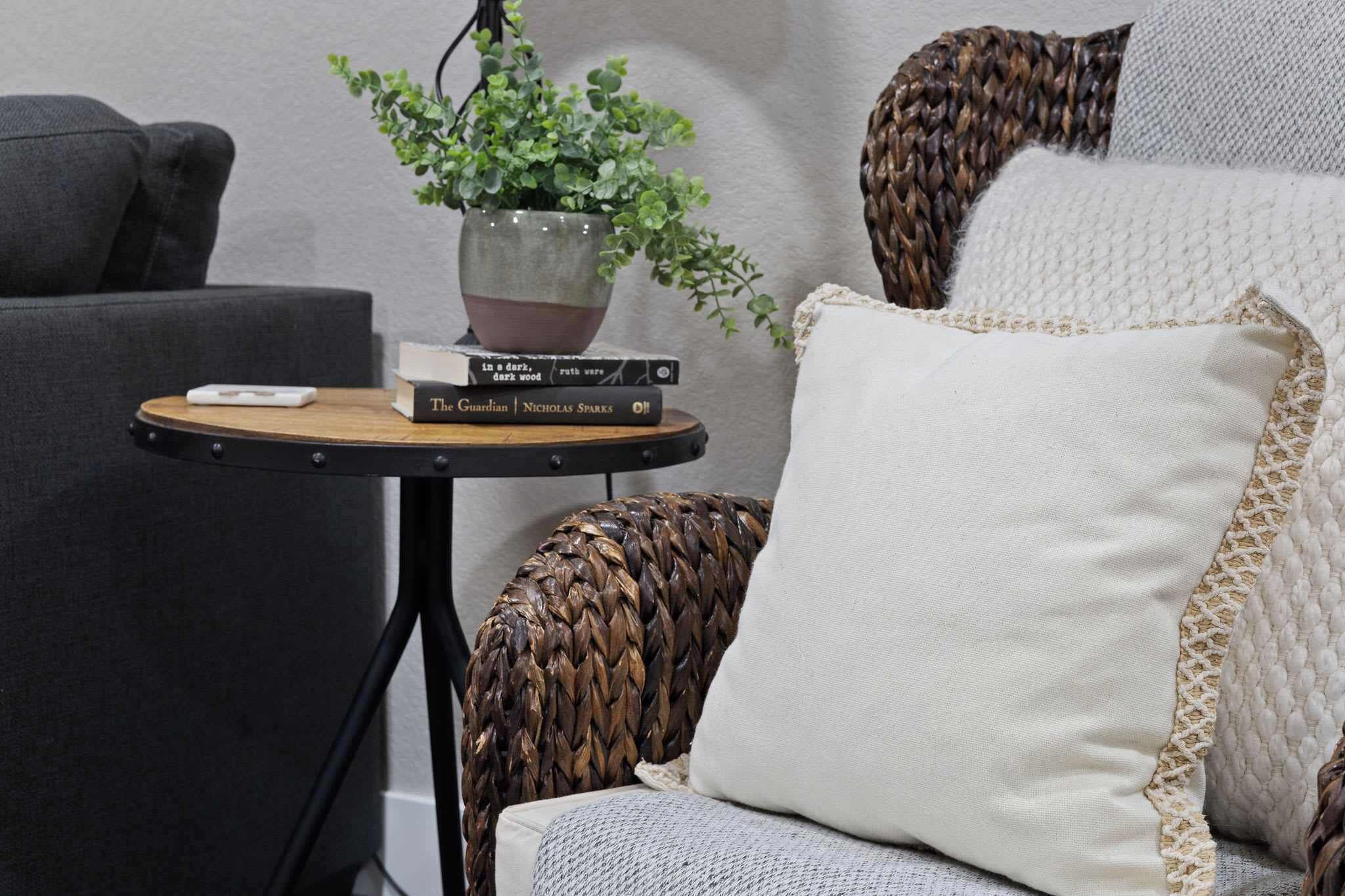 A cozy seating area with a woven chair, a small round table with a plant and books, and a white cushion.