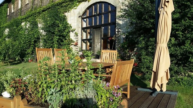 Una terraza de madera con una mesa y sillas, rodeada de abundante vegetación y un edificio de piedra con un marco de ventana negro.