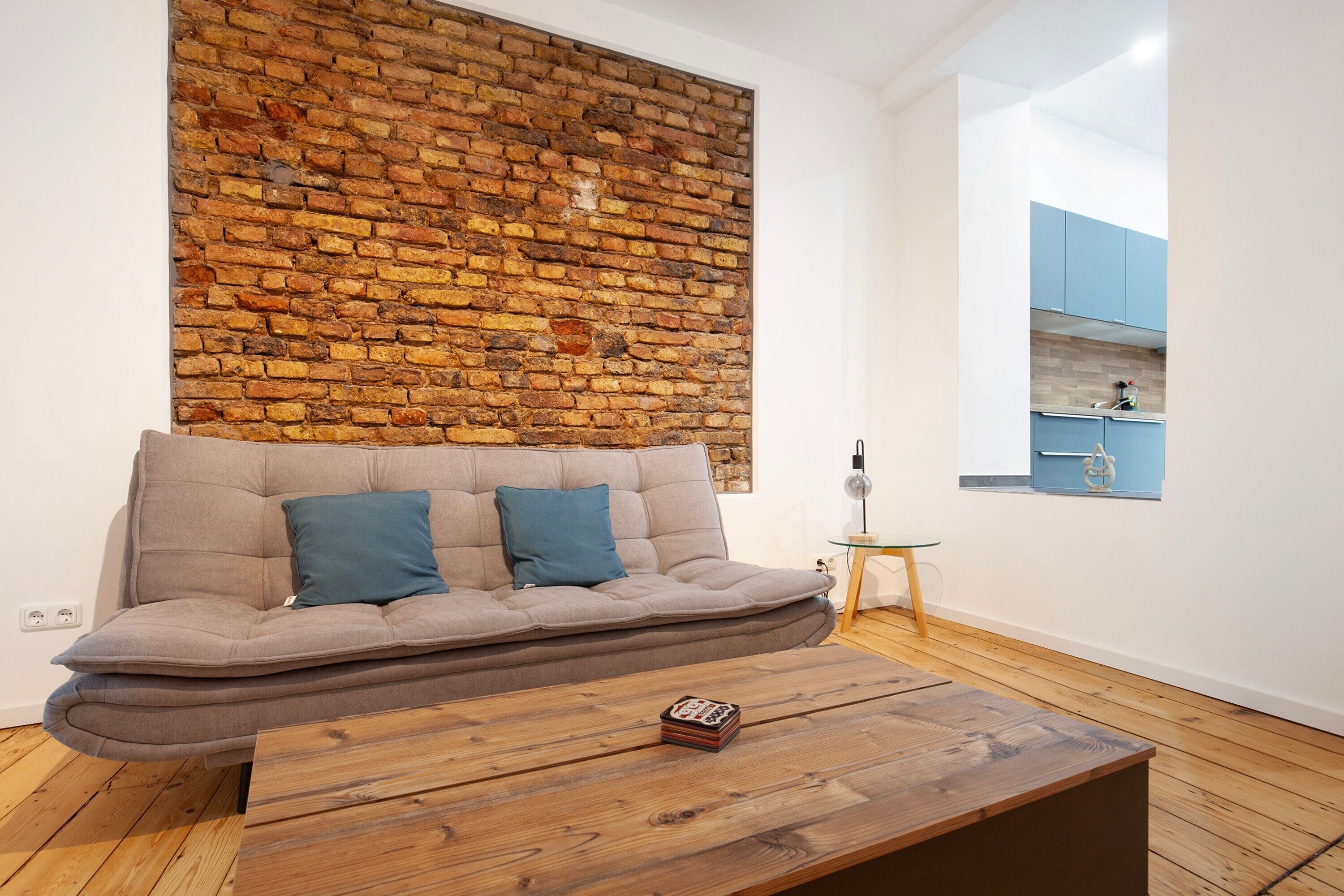 Un salon moderne avec un mur en briques, un canapé avec des coussins, une table basse en bois et une vue sur un espace cuisine.