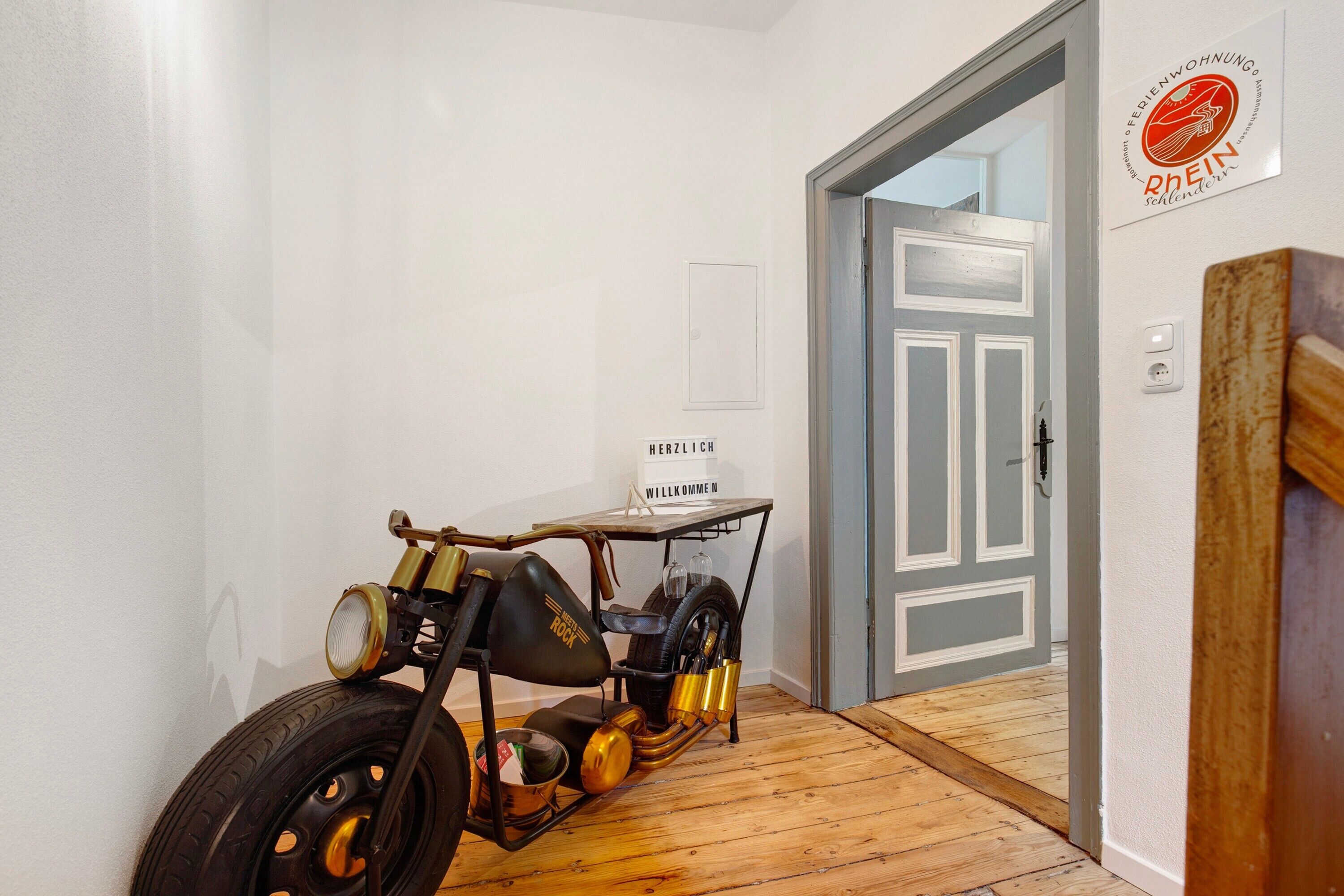 Ein altes Motorrad steht in einer Diele mit Holzboden und einer Tür zu einem weiteren Zimmer.