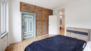 Een slaapkamer met houten vloer, een bed met donkere deken, een garderobe, een deur met blauwe omlijsting en een bakstenen muur.