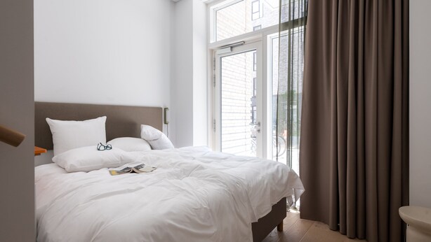 A bedroom with a large bed, a window with curtains, and a view of buildings outside. - Beach Gardens 46 by Daniel&Jacob's (København)