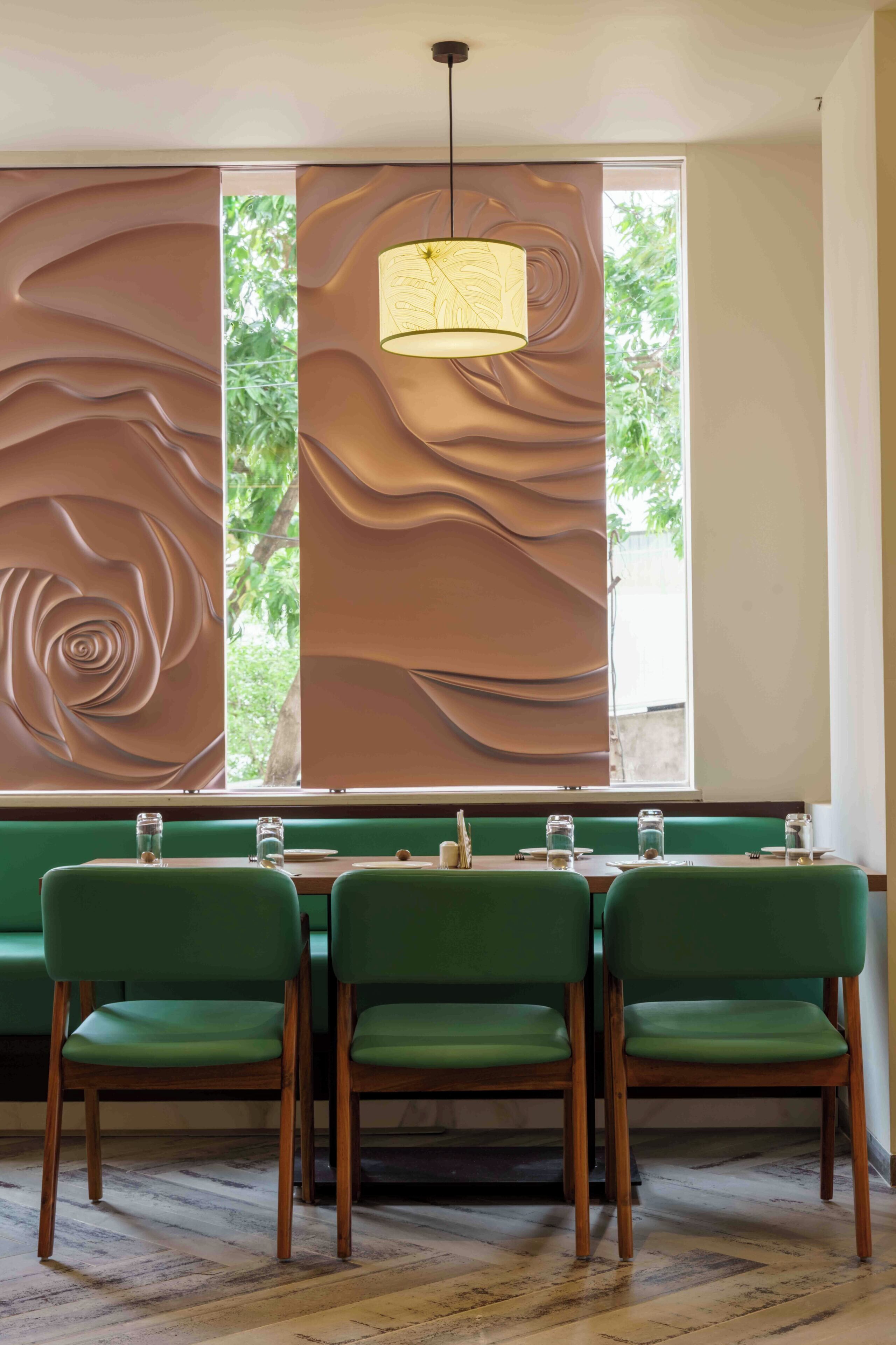 A dining area with a table set for three, green upholstered chairs, a hanging light fixture, and a decorative wall panel.