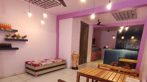 Una habitación con un banco de madera, una cama con colcha de cuadros rojos y blancos, un ventilador de techo y un estante en la pared con botellas y flores.