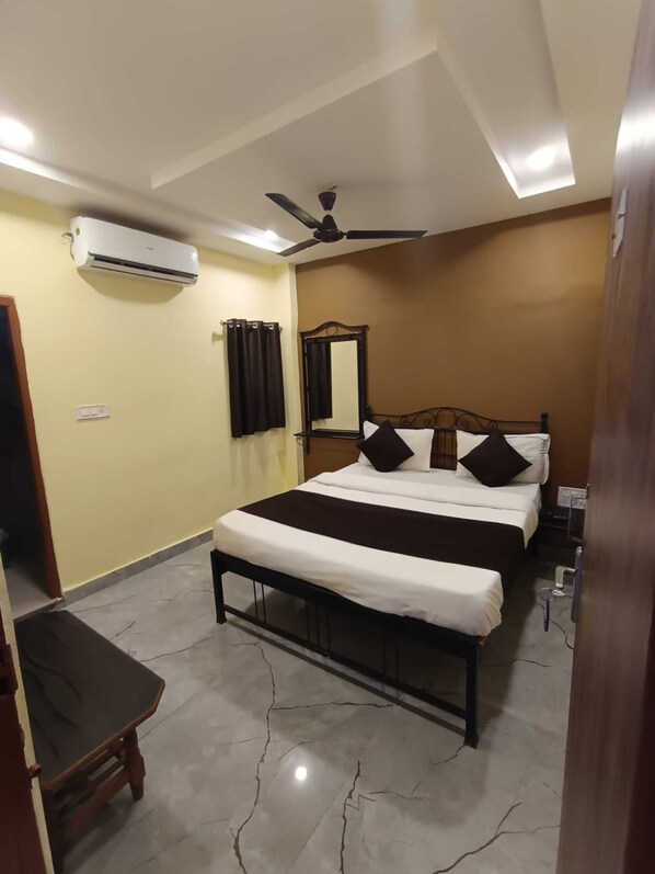 A bedroom with a bed, a ceiling fan, an air conditioning unit, a mirror, and a small wooden chair. - Hotel Mann (Indore)