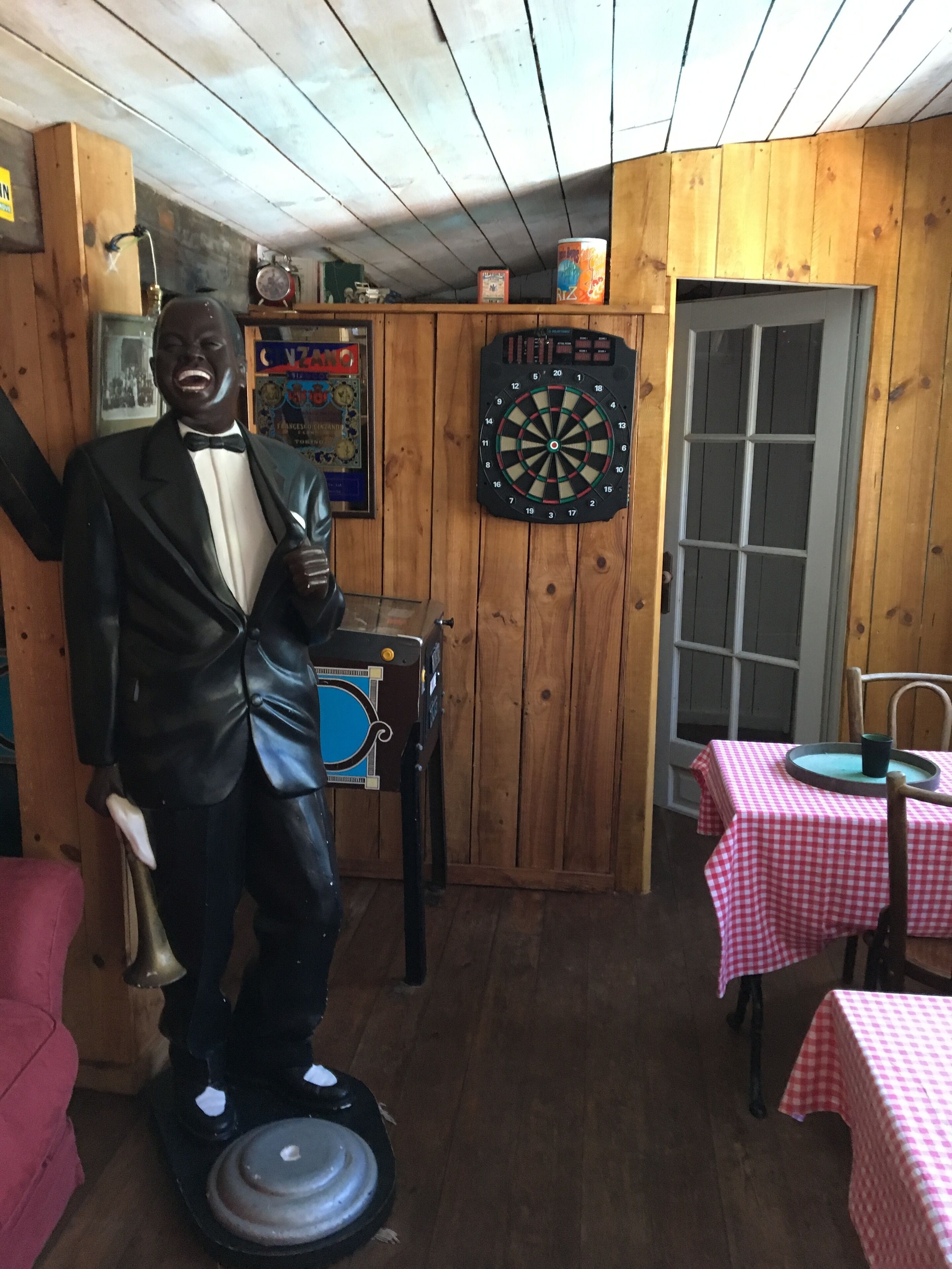 Une pièce aux murs en bois, avec un jeu de fléchettes, une statue, une table recouverte d’une nappe à carreaux et une porte.