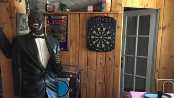 Une pièce aux murs en bois, avec un jeu de fléchettes, une statue, une table recouverte d’une nappe à carreaux et une porte.