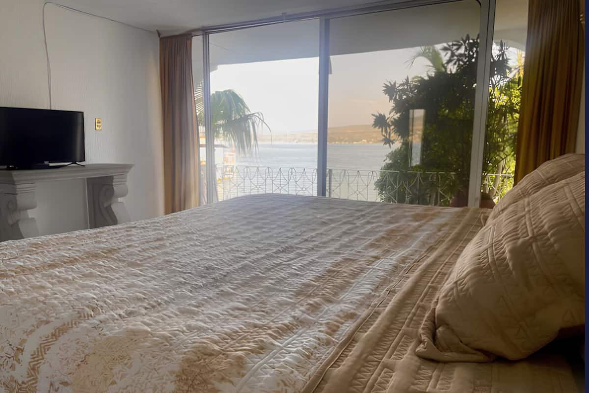 Een hotelkamer met een groot bed, een televisie op een standaard en uitzicht op de zee door grote ramen.