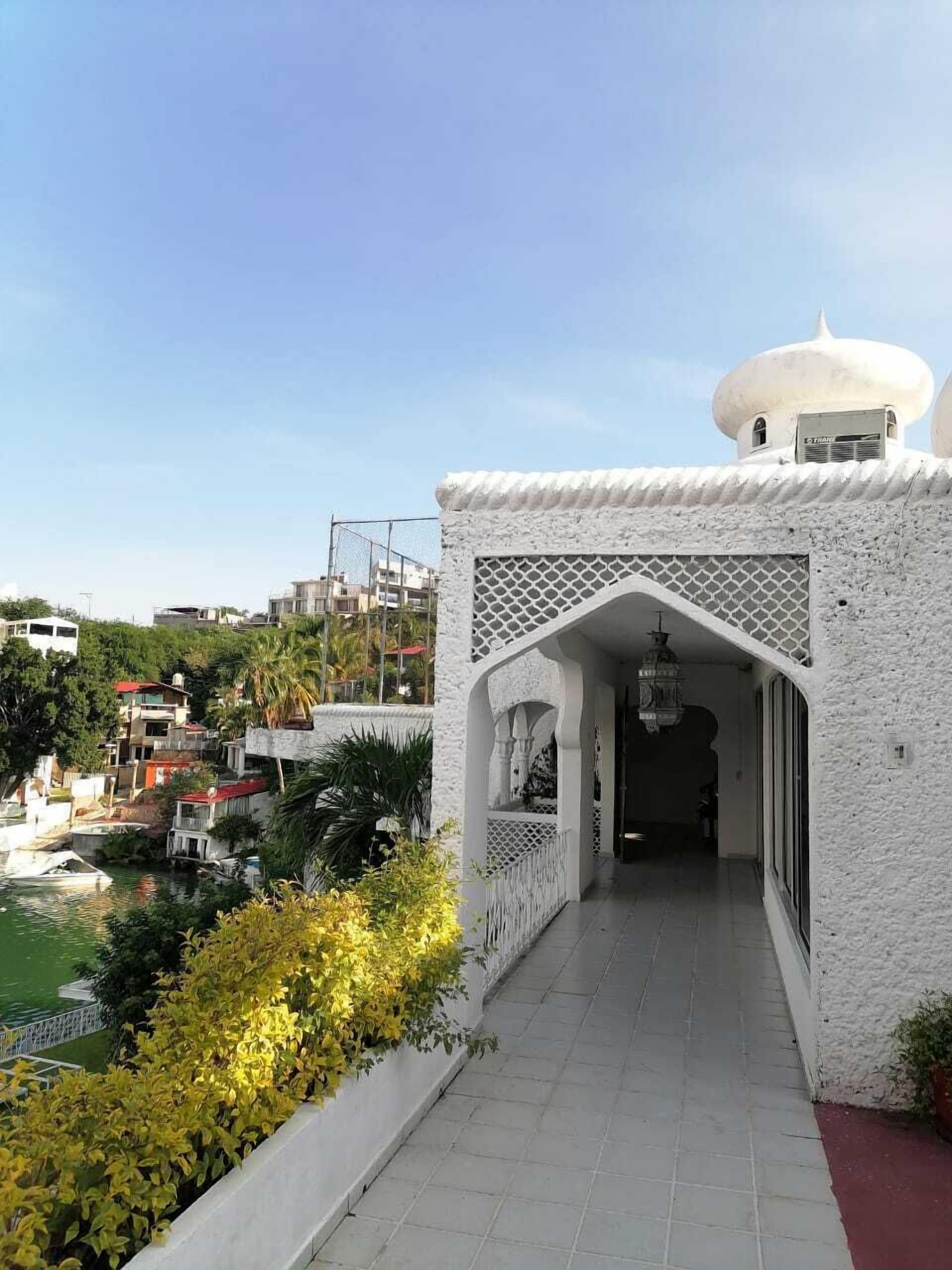 A white building with a covered walkway leading to a door, surrounded by greenery and a body of water.