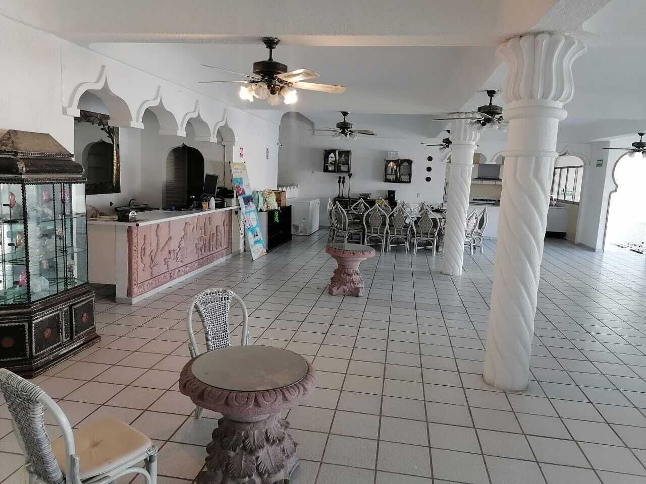 Un hall d’entrée spacieux avec un sol carrelé de blanc, des colonnes blanches et des ventilateurs de plafond. On y trouve plusieurs tables et chaises, un comptoir d’accueil et une vitrine décorative.