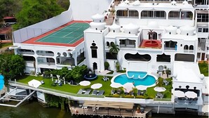 Um edifício grande e branco, com vários andares, possui quadra de tênis no terraço, piscina e um píer com barco.