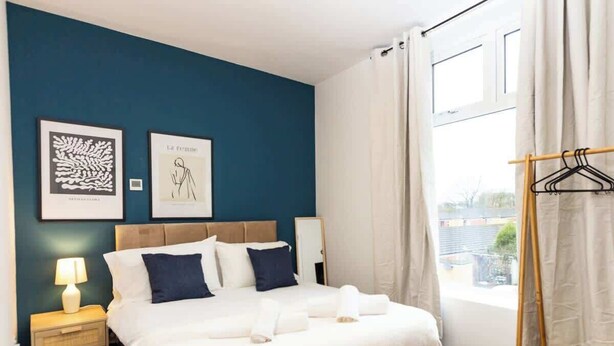 Une chambre d’hôtel comprenant un lit, une table de chevet, une lampe, une fenêtre avec des rideaux et deux œuvres d’art encadrées accrochées au mur.