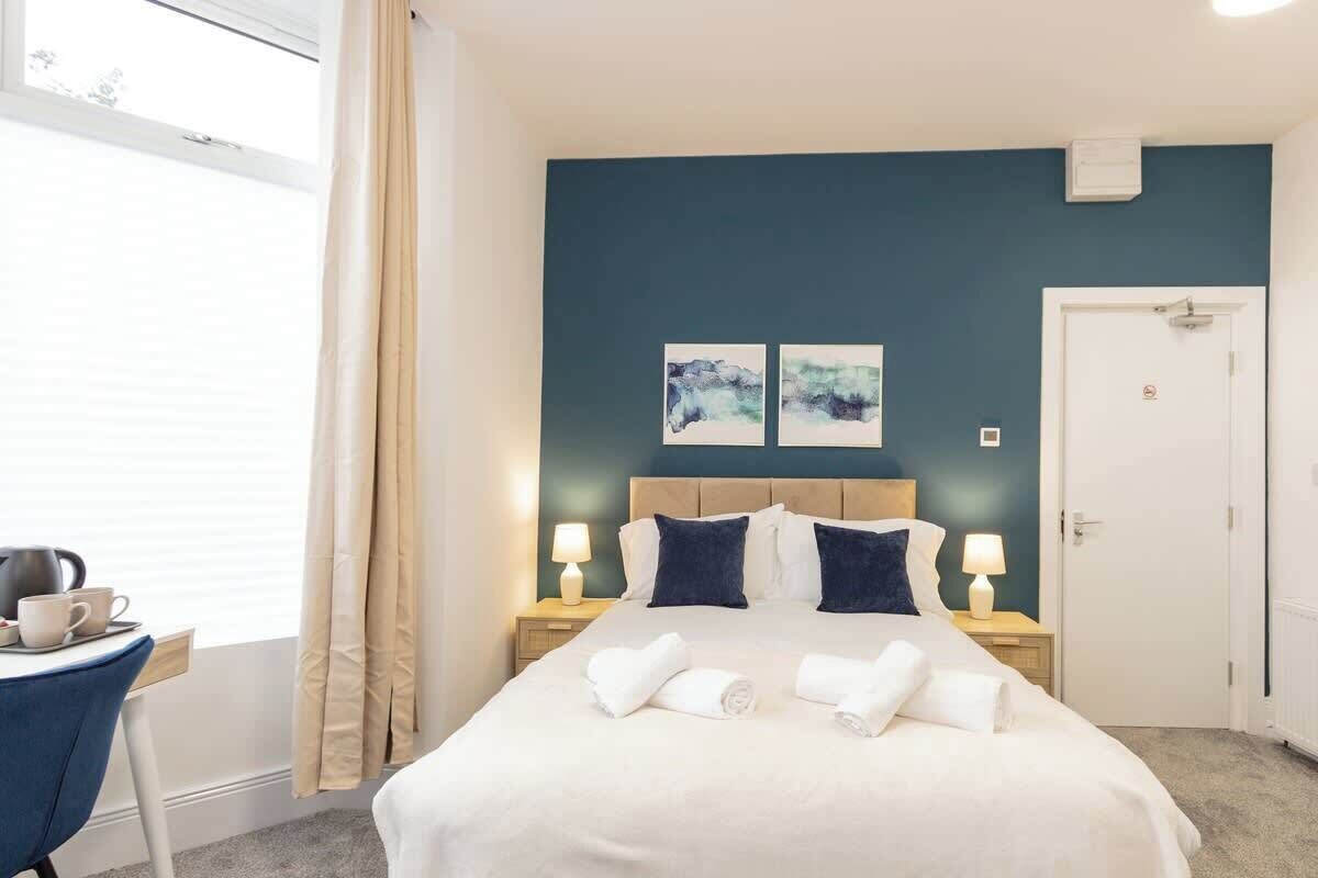 A hotel room with a bed, two bedside lamps, a window with curtains, a desk with a kettle, and two wall art pieces.