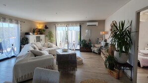 Ein geräumiges Wohnzimmer mit einer großen Couch, einem Couchtisch und einem Fernseher. Mehrere Zimmerpflanzen und eine Buddha-Statue sind vorhanden. Das Zimmer verfügt über große Fenster mit durchsichtigen Vorhängen und einer Klimaanlage an der Wand.
