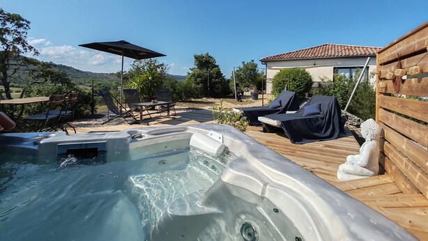 Ein Whirlpoolbereich mit Liegestühlen und einem Sonnenschirm, umgeben von einer Holzdielen-Terrasse und einem Haus mit einem Ziegeldach.