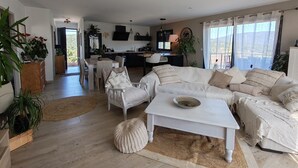 Ein geräumiger Wohnbereich mit einer weißen Sofagarnitur, einem Couchtisch und einem Esstisch. Im Hintergrund befindet sich eine Küche mit modernen Geräten und einem großen Fenster.