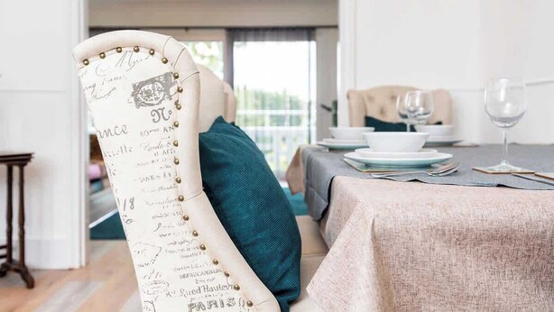 A dining area with a table set for two, a chair with decorative writing, and a view of the outdoors. - 3 BR Chic Home with Balcony & Free Parking! (Leeds)