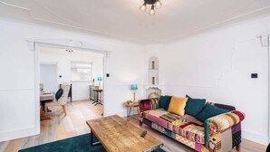 A living room with a wooden coffee table, a colorful sofa, and a teal rug. - 3 BR Chic Home with Balcony & Free Parking! (Leeds)