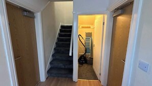En hall med ett mattbeklätt golv, en trädörr till vänster, en trappa med ett ledstång och en dammsugare i hörnet.