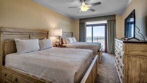 Habitación de hotel con dos camas, ventilador de techo, cómoda y un ventanal grande con cortinas.