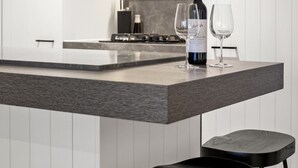 Une cuisine moderne avec un îlot central, deux tabourets de bar noirs, une bouteille de vin et deux verres à vin.