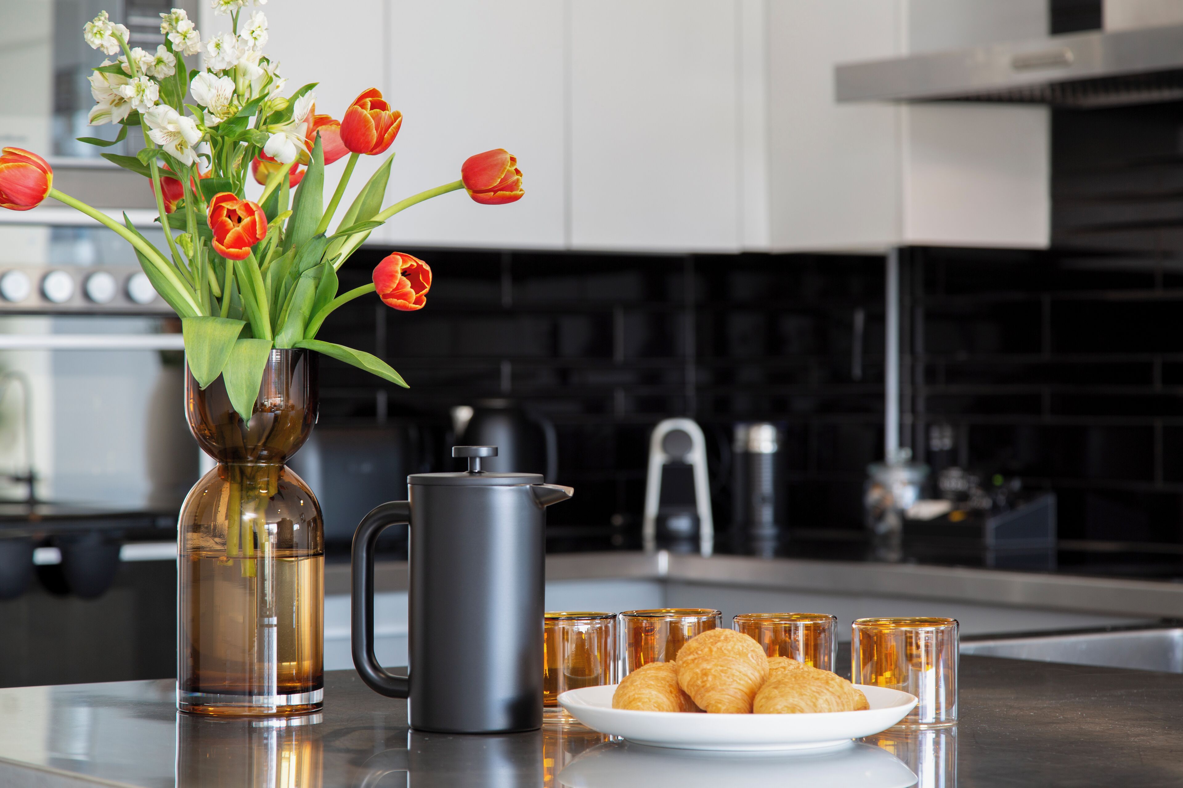 A kitchen counter with a vase of tulips, a coffee pot, and a plate of croissants.