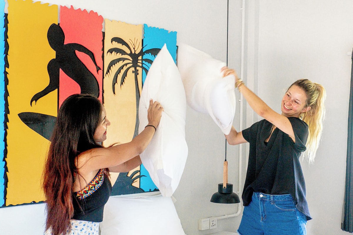 Two people holding a large white pillow in a room with a colorful mural on the wall and a window with curtains.